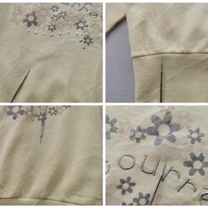 May include: A light yellow sweatshirt with a floral design and the word "ourre" embroidered on the front. The floral design includes gray and pink flowers with silver studs. The sweatshirt has a ribbed hem and cuffs.