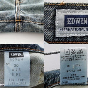 May include: Close-up of blue denim jeans with visible stitching and labels. The labels display the brand "EDWIN" and "INTERNATIONAL BASIC", along with size details: waist 30 inches, waist 76 cm, and hips 93 cm. Additional tags provide care instructions.