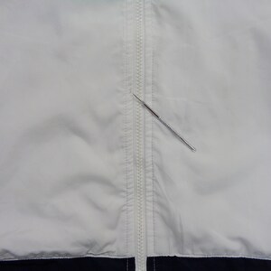 May include: A white jacket with a full-length zipper down the front. The jacket has navy blue accents at the bottom and on the sides. A silver metal tool is placed diagonally across the zipper.
