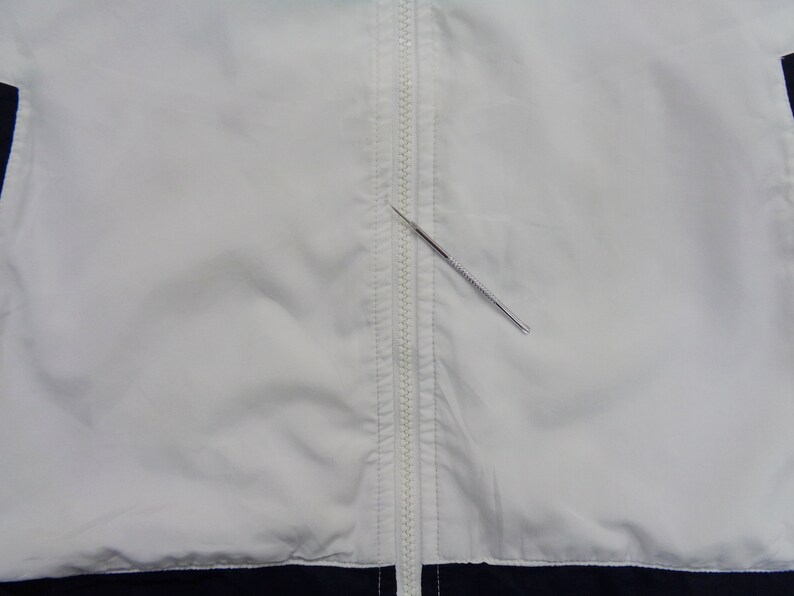 May include: A white jacket with a full-length zipper down the front. The jacket has navy blue accents at the bottom and on the sides. A silver metal tool is placed diagonally across the zipper.