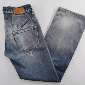 May include: Faded blue denim jeans with a distressed appearance. They feature a brown leather patch and a red tag. The jeans have a classic five-pocket design and a straight leg cut.