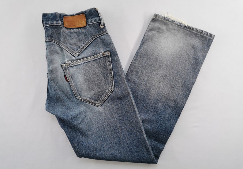 May include: Faded blue denim jeans with a distressed appearance. They feature a brown leather patch and a red tag. The jeans have a classic five-pocket design and a straight leg cut.