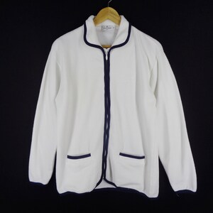 May include: White zip-up jacket with navy blue trim and two front pockets. The jacket is hanging on a wooden hanger against a black background. The jacket has long sleeves and a collar.