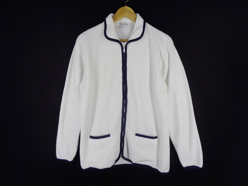 May include: White zip-up jacket with navy blue trim and two front pockets. The jacket is hanging on a wooden hanger against a black background. The jacket has long sleeves and a collar.