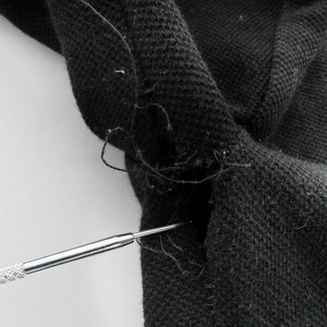May include: Close-up of black fabric with a visible tear and loose threads. A silver tool is inserted into the tear, highlighting the damage. The fabric appears to be a knit or woven material.