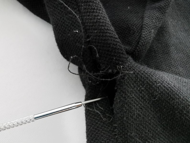 May include: Close-up of black fabric with a visible tear and loose threads. A silver tool is inserted into the tear, highlighting the damage. The fabric appears to be a knit or woven material.