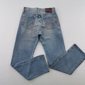May include: Light blue denim jeans with a classic five-pocket design. The jeans feature a faded wash, a brown leather patch on the back, and detailed stitching. The straight-leg cut offers a timeless style.