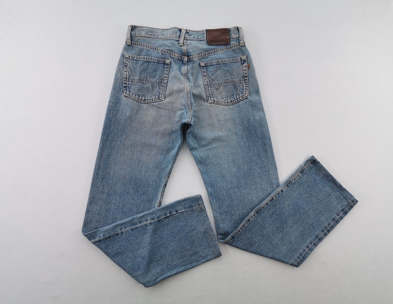 May include: Light blue denim jeans with a classic five-pocket design. The jeans feature a faded wash, a brown leather patch on the back, and detailed stitching. The straight-leg cut offers a timeless style.