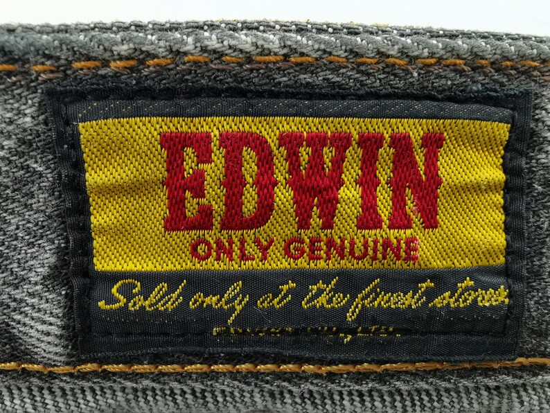 May include: Close-up of a denim label with the brand name "EDWIN" in red letters on a yellow background. The label also includes the text "ONLY GENUINE" and "Sold only at the finest stores."
