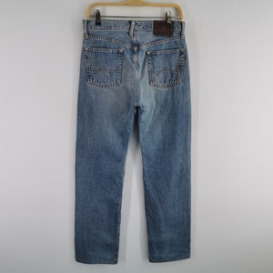 May include: Light blue denim jeans with a classic five-pocket design, visible back pockets, and a brown leather patch. The jeans are hanging on a wooden hanger against a plain white background.