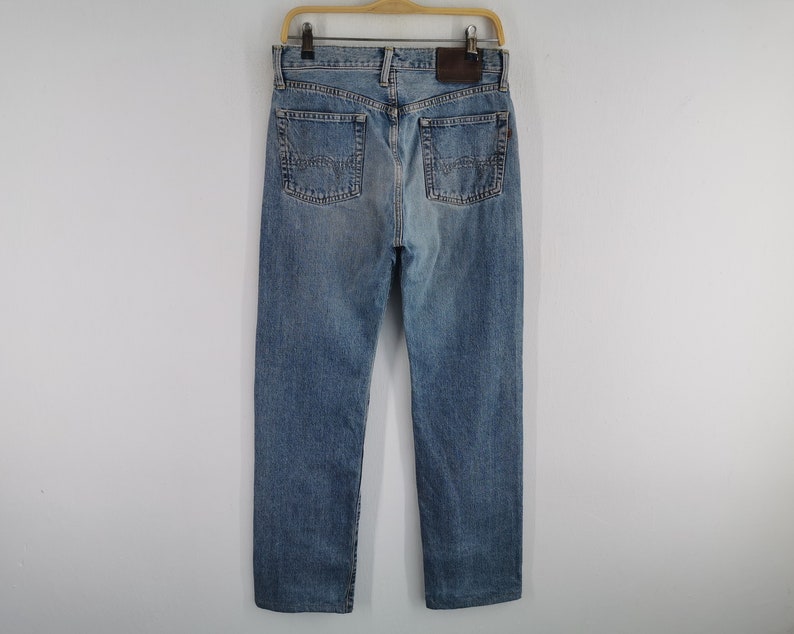 May include: Light blue denim jeans with a classic five-pocket design, visible back pockets, and a brown leather patch. The jeans are hanging on a wooden hanger against a plain white background.