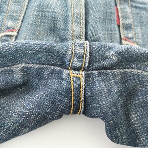 Pu&ograve; includere: Un paio di jeans in denim blu con cucitura del cavallo rinforzata e spesse impunture a contrasto color giallo oro. Il tessuto presenta un classico lavaggio indaco con una trama visibile, mentre piccoli accenti di filo rosso sono presenti.