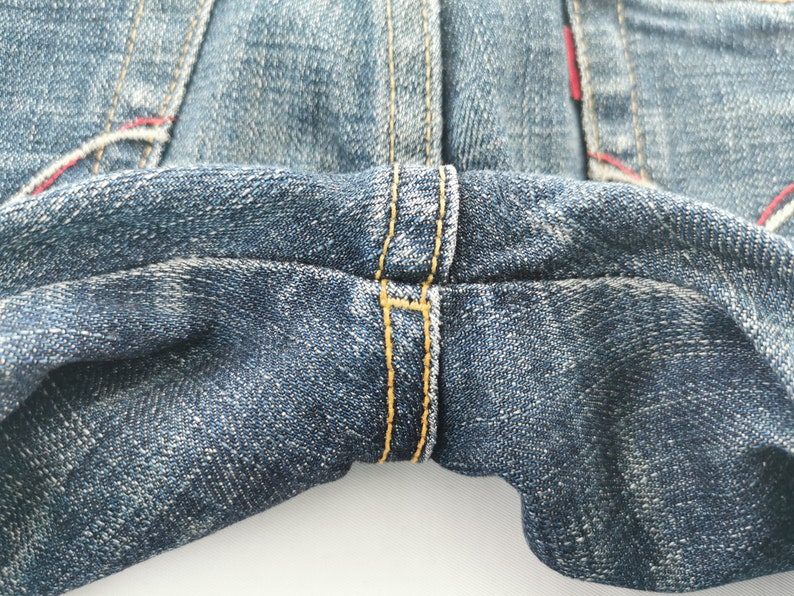 Pu&ograve; includere: Un paio di jeans in denim blu con cucitura del cavallo rinforzata e spesse impunture a contrasto color giallo oro. Il tessuto presenta un classico lavaggio indaco con una trama visibile, mentre piccoli accenti di filo rosso sono presenti.