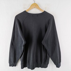 May include: A charcoal gray sweatshirt with long sleeves, hanging on a wooden hanger against a white wall. The crew neck sweatshirt has a faded appearance, suggesting it is vintage or has been washed many times.