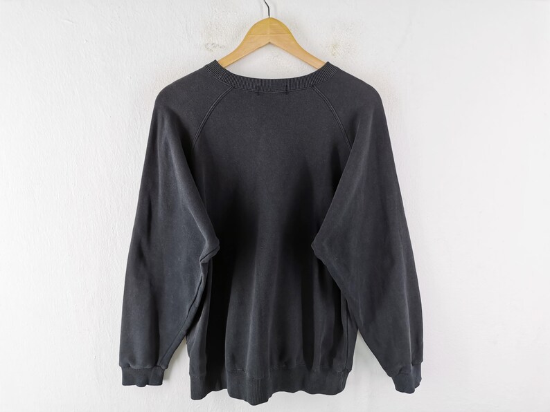 May include: A charcoal gray sweatshirt with long sleeves, hanging on a wooden hanger against a white wall. The crew neck sweatshirt has a faded appearance, suggesting it is vintage or has been washed many times.