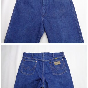 May include: Blue denim jeans with a classic five-pocket design. The jeans feature a button and zipper closure, with copper-colored rivets and a leather patch on the back pocket. The Wrangler logo is visible on the patch.