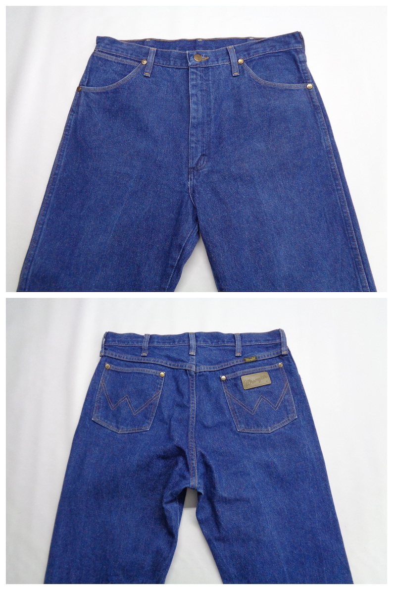 May include: Blue denim jeans with a classic five-pocket design. The jeans feature a button and zipper closure, with copper-colored rivets and a leather patch on the back pocket. The Wrangler logo is visible on the patch.