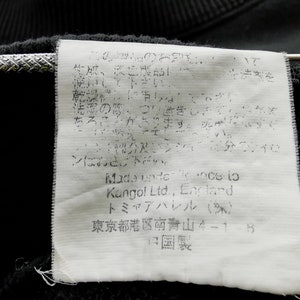 May include: Close-up of a black garment tag with Japanese text and English text. The tag includes the brand name "Kangol Ltd., England" and other Japanese text. The tag is attached to a black fabric.