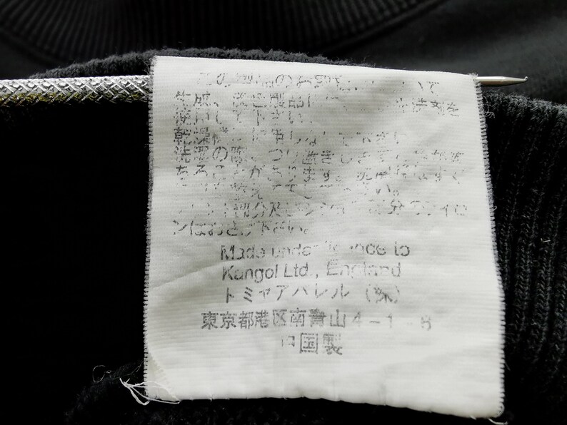 May include: Close-up of a black garment tag with Japanese text and English text. The tag includes the brand name "Kangol Ltd., England" and other Japanese text. The tag is attached to a black fabric.