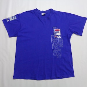 May include: A royal blue t-shirt with the Fila logo on the front and left sleeve. The logo is white with red and blue accents. The shirt is made of a soft material and has short sleeves.