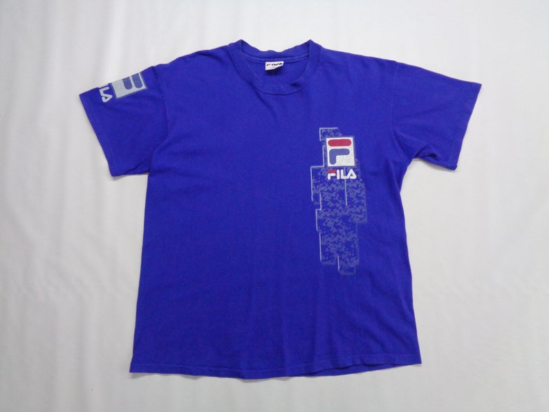 May include: A royal blue t-shirt with the Fila logo on the front and left sleeve. The logo is white with red and blue accents. The shirt is made of a soft material and has short sleeves.