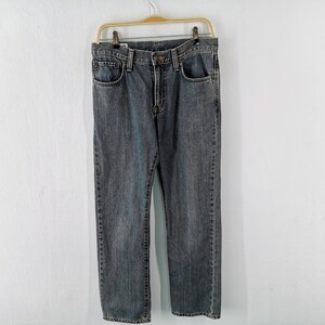 May include: A pair of blue denim jeans hanging on a gold-colored hanger against a white wall. The jeans have a classic five-pocket design and a straight leg fit. The denim has a faded, worn look.