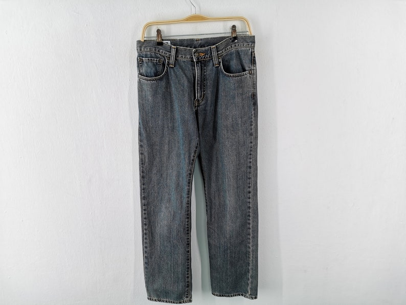 May include: A pair of blue denim jeans hanging on a gold-colored hanger against a white wall. The jeans have a classic five-pocket design and a straight leg fit. The denim has a faded, worn look.