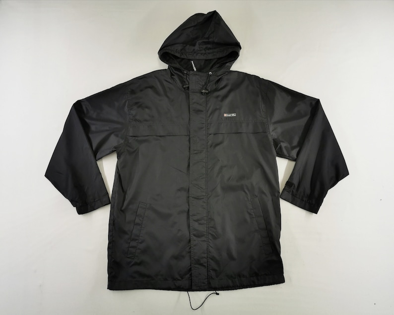 May include: A black hooded rain jacket with a drawstring hood and a small logo on the chest. The jacket has long sleeves and a front zipper closure. The material appears to be a lightweight, water-resistant fabric, suitable for wet weather.