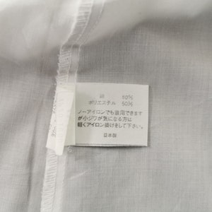 May include: Close-up of a white fabric with a sewn seam and a rectangular tag. The tag has Japanese text and reads "50% cotton, 50% polyester". Additional text advises light ironing if needed.