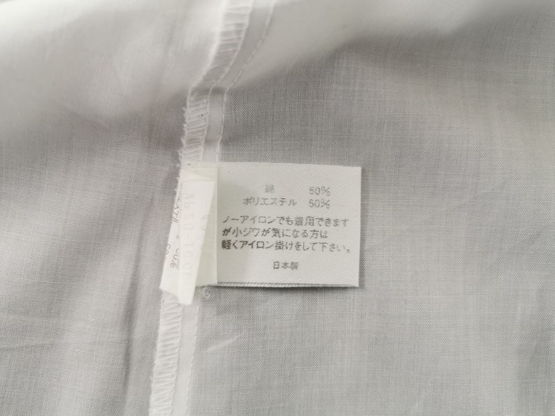 May include: Close-up of a white fabric with a sewn seam and a rectangular tag. The tag has Japanese text and reads "50% cotton, 50% polyester". Additional text advises light ironing if needed.
