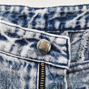 May include: Close-up of acid wash denim jeans with a silver button and a brass zipper. The button is embossed with text. The denim has a blue and white pattern.