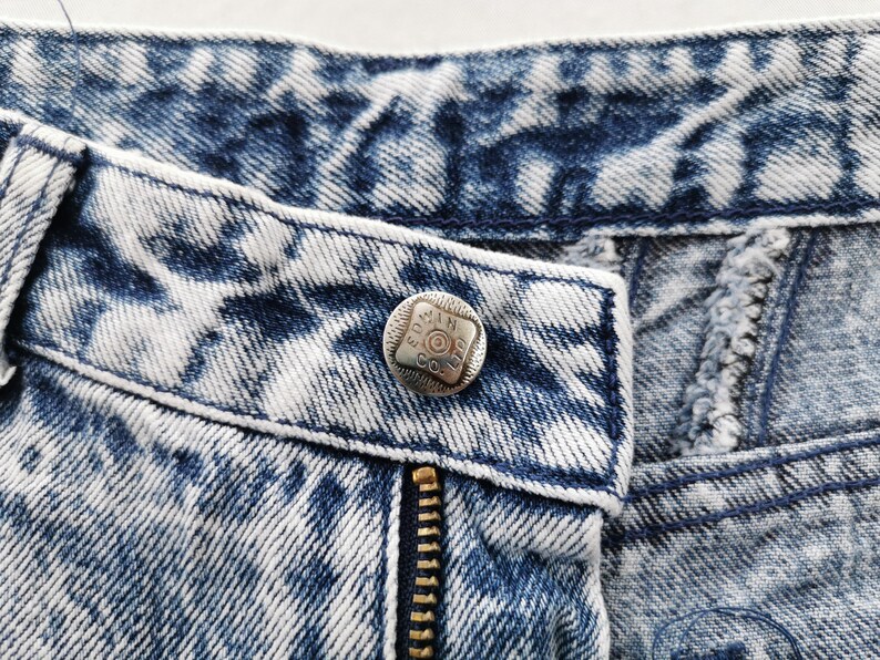 May include: Close-up of acid wash denim jeans with a silver button and a brass zipper. The button is embossed with text. The denim has a blue and white pattern.