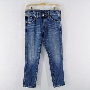 May include: Blue denim jeans hanging on a wooden hanger against a white wall. The jeans have a classic five-pocket design, a button and zipper closure, and a straight leg fit. The denim has a faded, worn look.