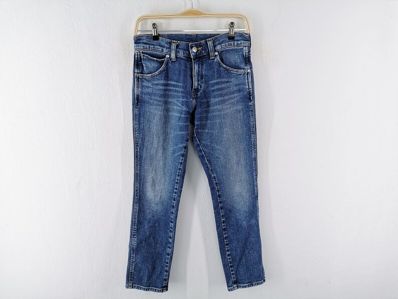 May include: Blue denim jeans hanging on a wooden hanger against a white wall. The jeans have a classic five-pocket design, a button and zipper closure, and a straight leg fit. The denim has a faded, worn look.