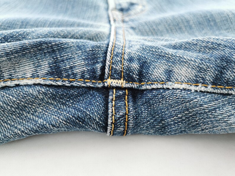 May include: Close-up of blue denim jeans with visible stitching. The jeans have a classic, worn look with yellow thread detailing along the seams. The fabric shows a textured pattern, highlighting the denim's weave.