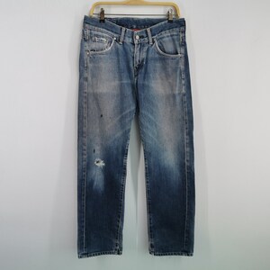 May include: Faded blue denim jeans with a distressed appearance, featuring a ripped knee and paint splatters. The jeans are hanging on a wooden hanger against a plain white background. A classic, casual style.