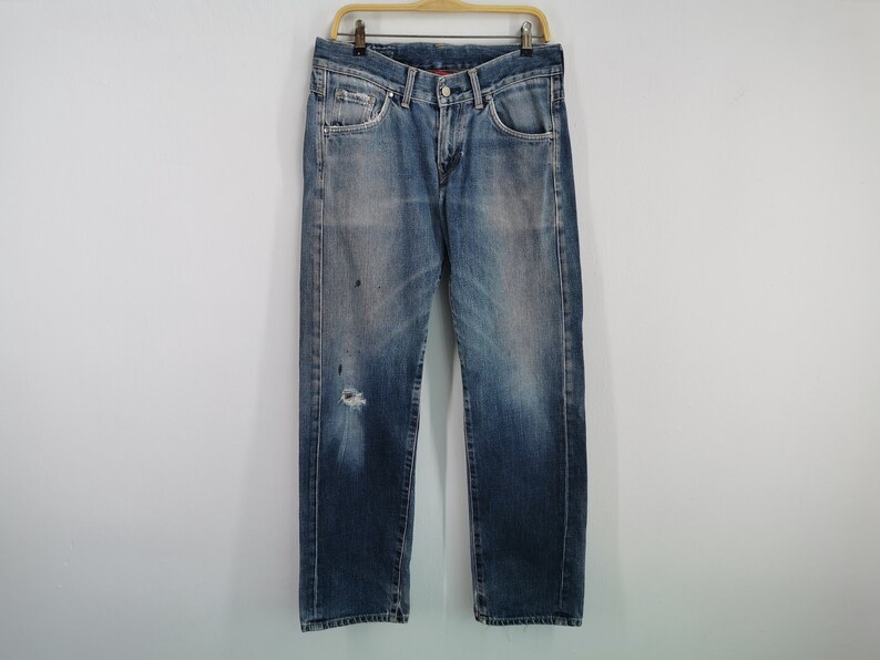 May include: Faded blue denim jeans with a distressed appearance, featuring a ripped knee and paint splatters. The jeans are hanging on a wooden hanger against a plain white background. A classic, casual style.