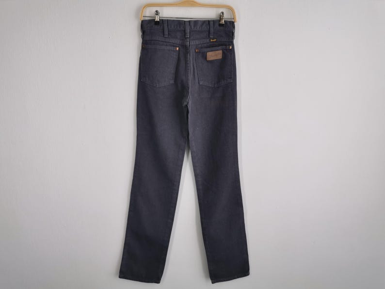 May include: Dark gray denim jeans hanging on a wooden hanger. The jeans have a classic five-pocket design, a brown leather patch on the back, and a straight leg cut. The jeans are made of a durable denim material.
