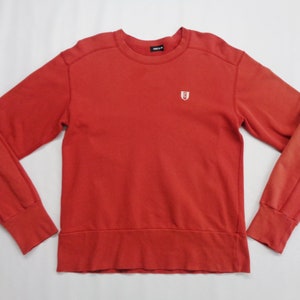 Puede incluir: Una sudadera roja con un pequeño emblema blanco en el pecho. La parte superior de manga larga tiene cuello redondo y puños y dobladillo acanalados. Los hombros y las mangas tienen un diseño único. El nombre de la marca es visible en el escote.