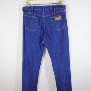 May include: Blue denim jeans with the Wrangler logo on the back pockets and a leather patch. The jeans are hanging on a wooden hanger against a white background. The stitching is a contrasting orange color.