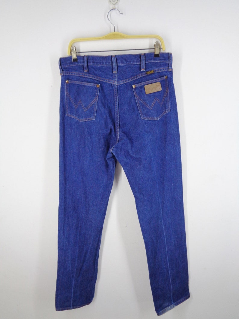 May include: Blue denim jeans with the Wrangler logo on the back pockets and a leather patch. The jeans are hanging on a wooden hanger against a white background. The stitching is a contrasting orange color.