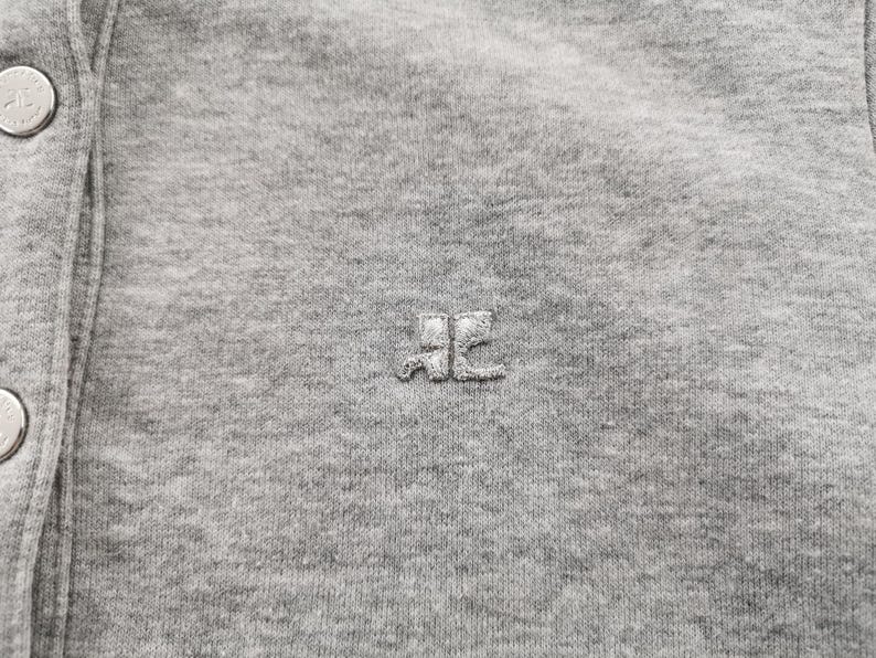 May include: Close-up of a gray, button-down garment. The fabric is a soft, textured knit. A small, embroidered logo is centered on the chest. Silver buttons are visible on the left side. The overall aesthetic is casual and comfortable.