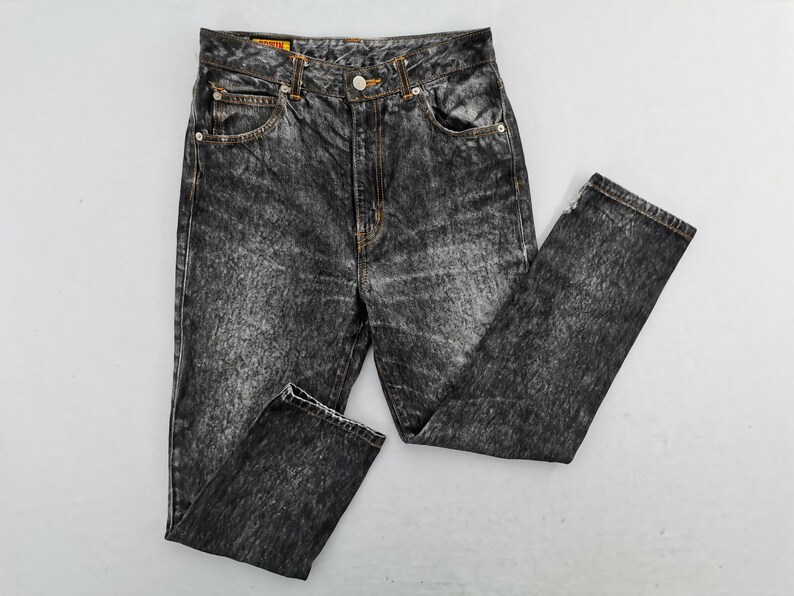 May include: Acid wash denim jeans with a classic five-pocket design. The jeans feature a button and zipper closure, belt loops, and a faded, textured appearance. The jeans are a dark gray color.