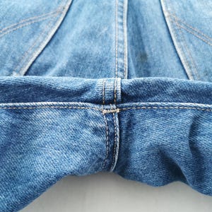 May include: Close-up of blue denim jeans, showcasing the back pockets and waistband. The stitching is visible in a contrasting thread, highlighting the seams and construction details. The fabric has a classic, worn look.