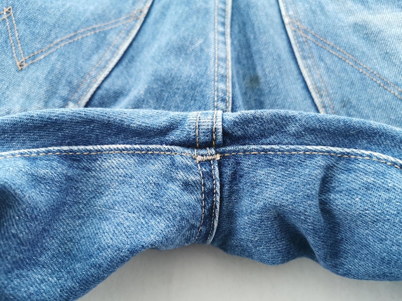 May include: Close-up of blue denim jeans, showcasing the back pockets and waistband. The stitching is visible in a contrasting thread, highlighting the seams and construction details. The fabric has a classic, worn look.