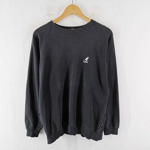 May include: A black sweatshirt with long sleeves, a crew neck, and a small white kangaroo logo on the chest. The sweatshirt is hanging on a wooden hanger against a white background. The Kangol logo is visible.