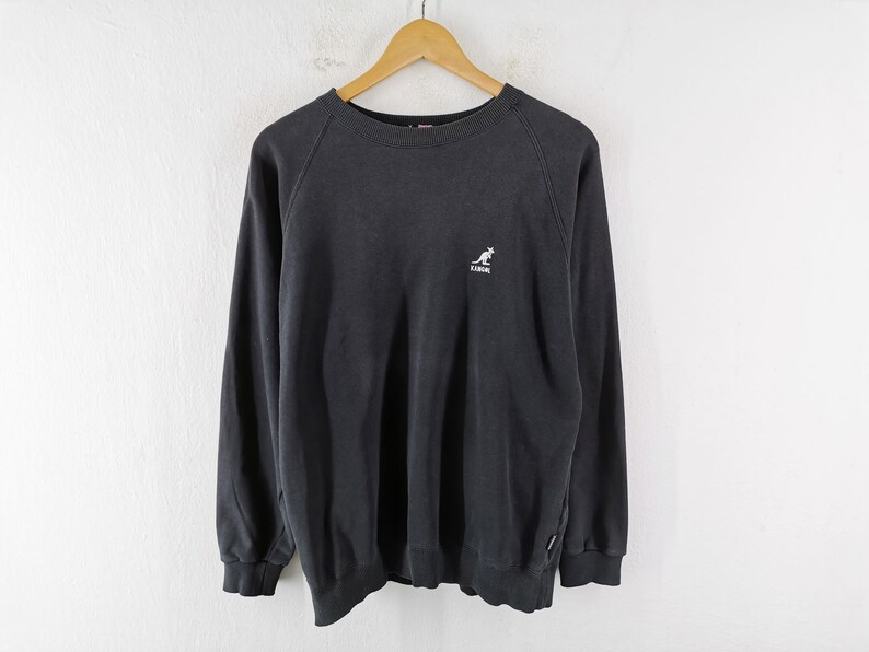 May include: A black sweatshirt with long sleeves, a crew neck, and a small white kangaroo logo on the chest. The sweatshirt is hanging on a wooden hanger against a white background. The Kangol logo is visible.