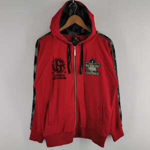 May include: Red zip-up hoodie with a hood and camouflage accents on the sleeves. The front features the text "USA Urban Division" and a patch that reads "BAD BOY USA". The hoodie is hanging on a wooden hanger.