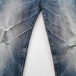 May include: Close-up of distressed blue denim jeans. The jeans have faded areas and two knee rips with frayed edges. The fabric shows wear and tear, with visible stitching and a light wash effect.