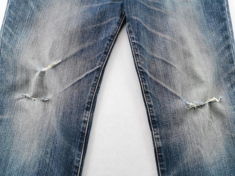 May include: Close-up of distressed blue denim jeans. The jeans have faded areas and two knee rips with frayed edges. The fabric shows wear and tear, with visible stitching and a light wash effect.